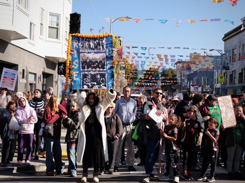 A crowd gathers in a street festival. A person holds up a sign covered in pictures and decorations. Colorful banners hang overhead, and people of all ages are present.