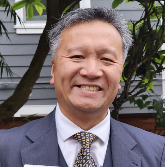 A person in a suit smiling outdoors, with a house and greenery in the background.