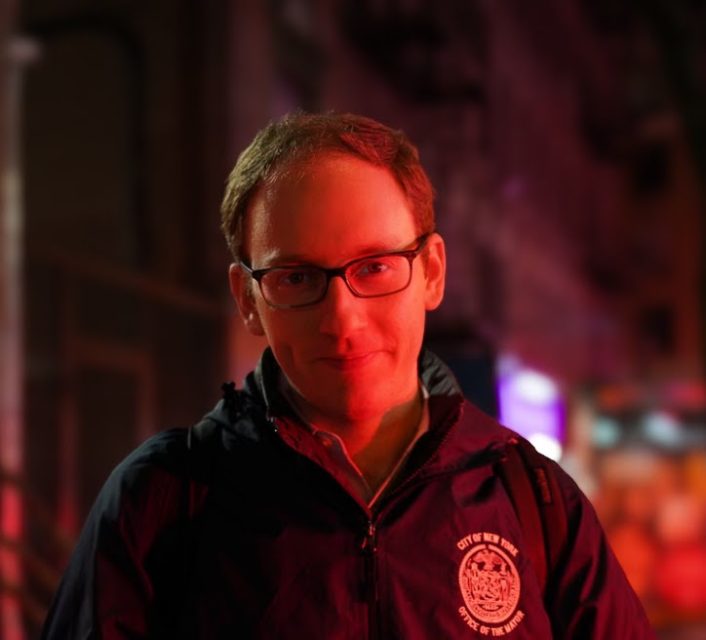 A person wearing glasses and a jacket with a city emblem stands in a dimly lit urban setting with a red tint.