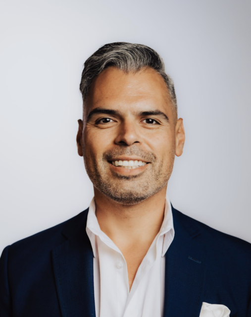 A man with short gray hair smiles, wearing a navy blazer and white shirt against a plain background.