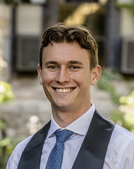 A person in a formal outfit smiles outdoors, with greenery and a building in the background.