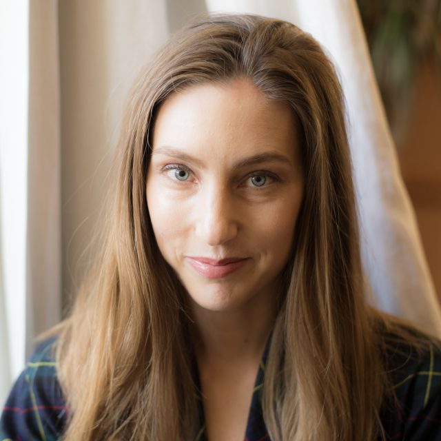 A woman with long brown hair sits in front of a window with light-colored curtains, wearing a dark plaid shirt.