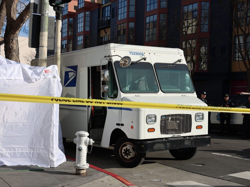A postal truck is stopped in a street with police tape and a white tent nearby. Buildings and officers are visible in the background.
