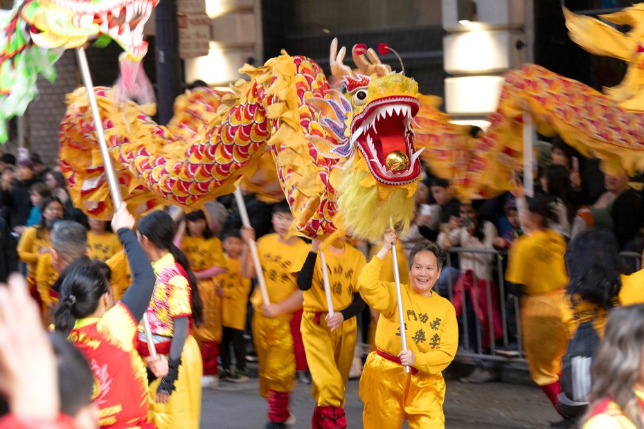Performers in yellow outfits carry a colorful dragon puppet in a lively street parade, with a crowd watching from the sidewalk.