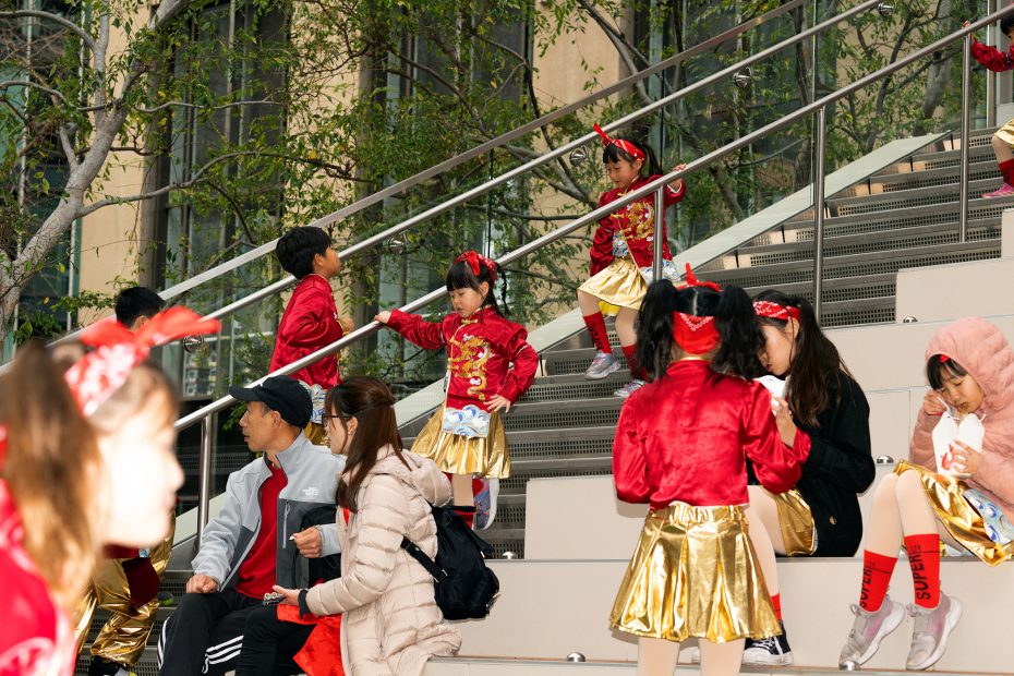 Children in coordinated red and gold outfits interact and rest on outdoor steps. Some wear jackets. The scene includes other people seated nearby, with trees in the background.