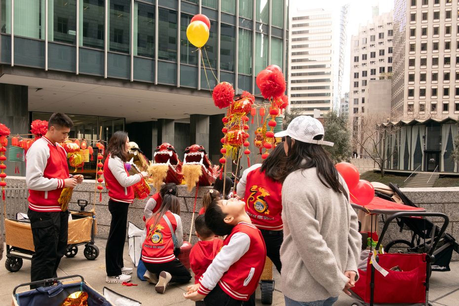 A group of people in red jackets prepares for a celebration outside, with decorative red lanterns and dragon costumes. Buildings are in the background.