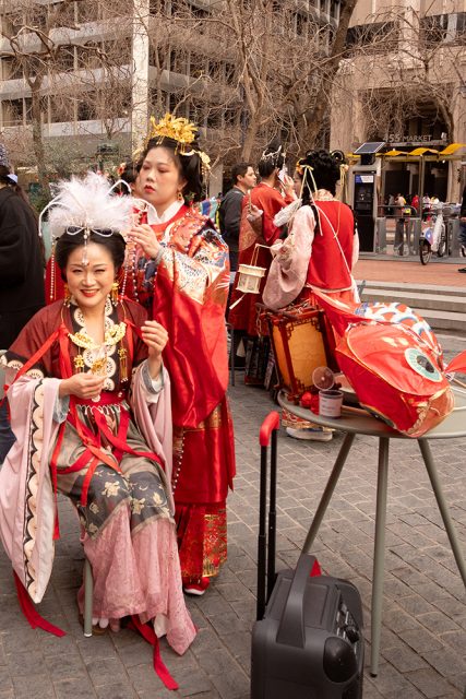 Two women in traditional attire with ornate hairpieces. One is seated, holding a bowl, while the other adjusts her hair. A suitcase and table with a drink are nearby. Urban background.