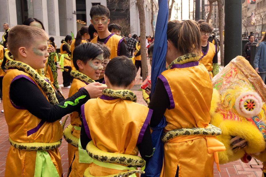 Group of children in yellow and purple costumes with snake patterns, discussing near a lion dance head on a city street.