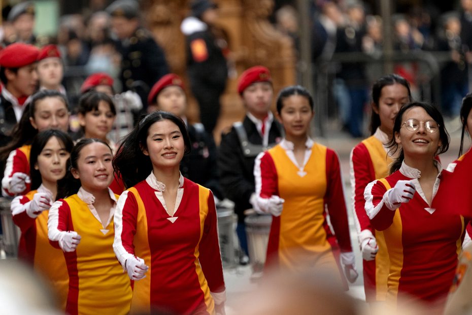Group of people in red and yellow uniforms marching in a parade, with onlookers and some members in red berets in the background.