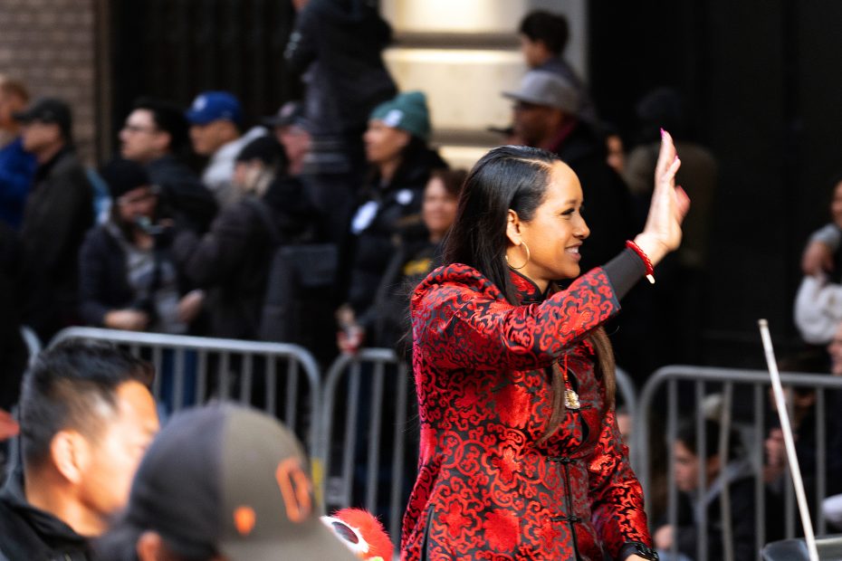 Person in a red patterned coat waves while standing in front of a crowd gathered behind a barrier.