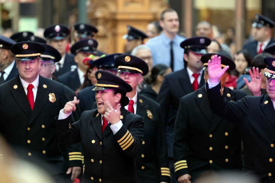 A group of uniformed individuals in black coats and red ties walk in a crowd. Some are gesturing with their hands.