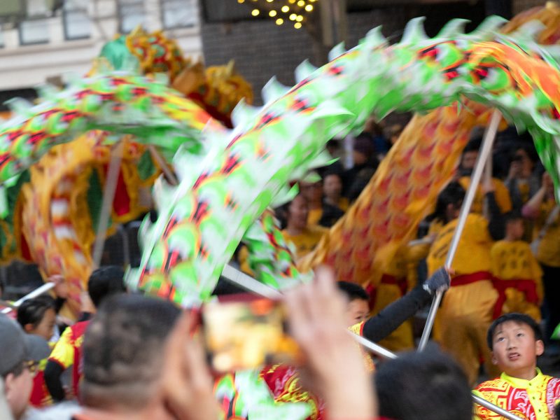 A group of performers in colorful dragon costumes participate in a lively celebration, with a crowd watching and capturing the moment.