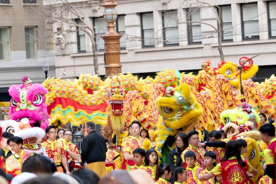 A group of people in red and yellow costumes perform a traditional dragon dance on a city street, with vibrant dragon figures above them.