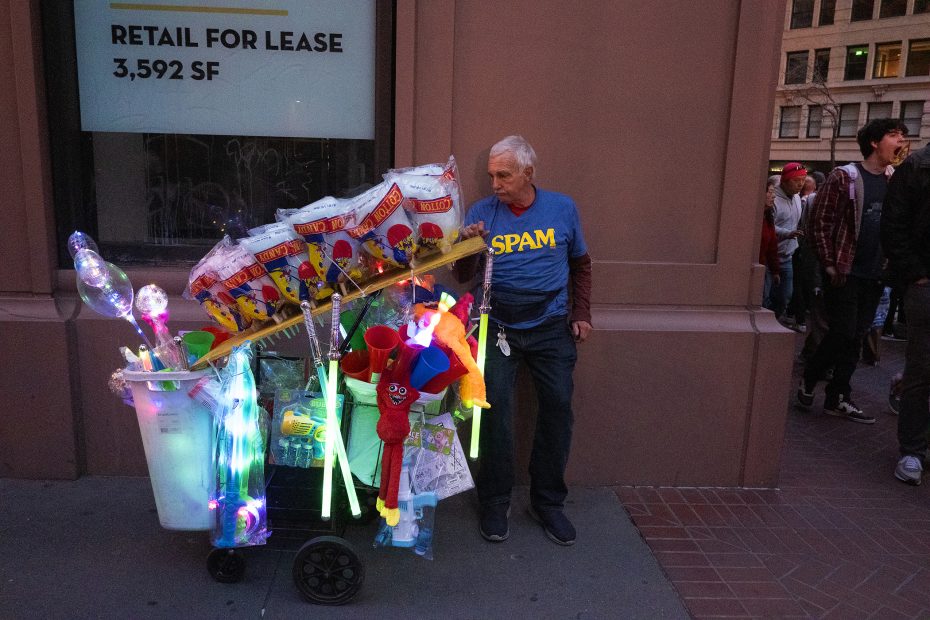 A vendor with a cart filled with light-up toys, balloons, and snacks stands on a city sidewalk, wearing a shirt with "SPAM" written on it.