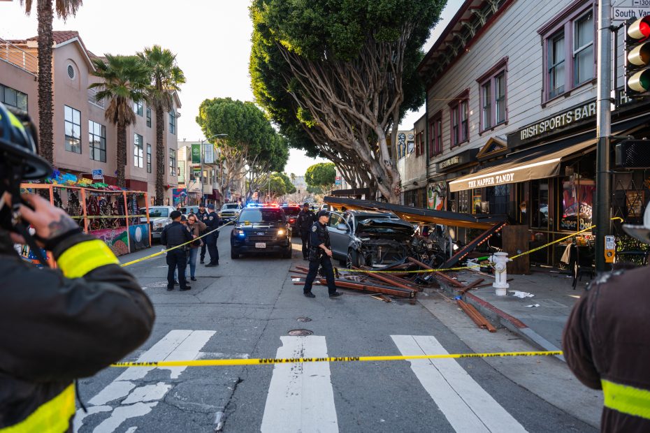 A car crash scene shows a vehicle damaged by a collapsed structure in an urban area, possibly linked to an intense police chase. Police officers and firefighters are present, with caution tape cordoning off the site, ensuring safety amid the chaos.