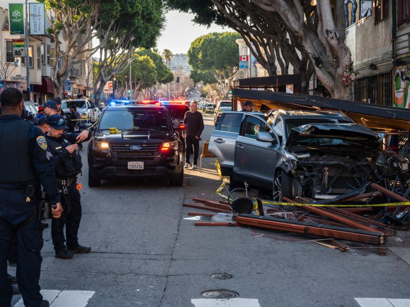 Police officers are at the scene of a vehicle crash, following an intense police chase, where a car has collided with a wooden structure on an urban street. The area is surrounded by debris and curious onlookers.