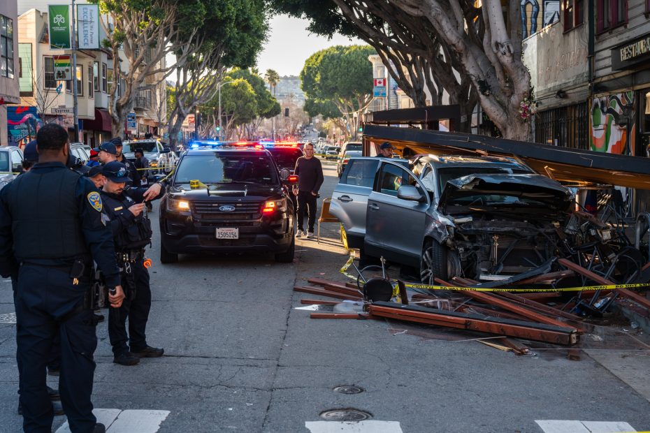 Police officers are at the scene of a vehicle crash, following an intense police chase, where a car has collided with a wooden structure on an urban street. The area is surrounded by debris and curious onlookers.