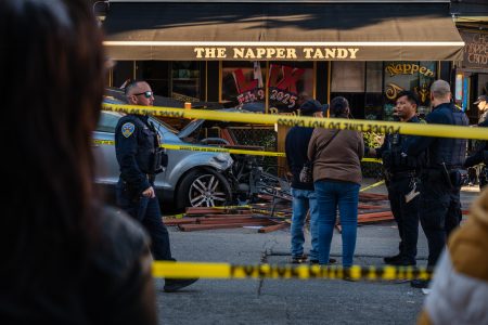 Police officers and bystanders at a car crash scene outside The Napper Tandy pub. Yellow caution tape blocks the area.