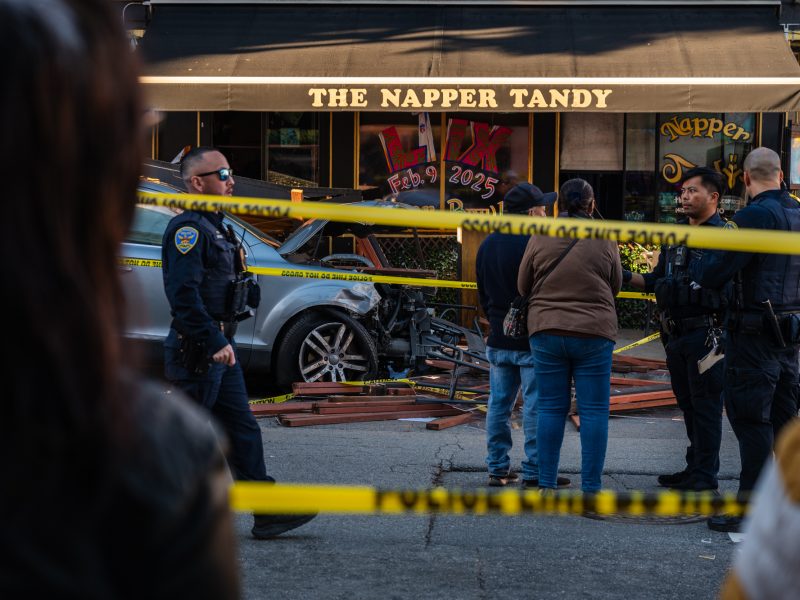 Police officers and bystanders at a car crash scene outside The Napper Tandy pub. Yellow caution tape blocks the area.