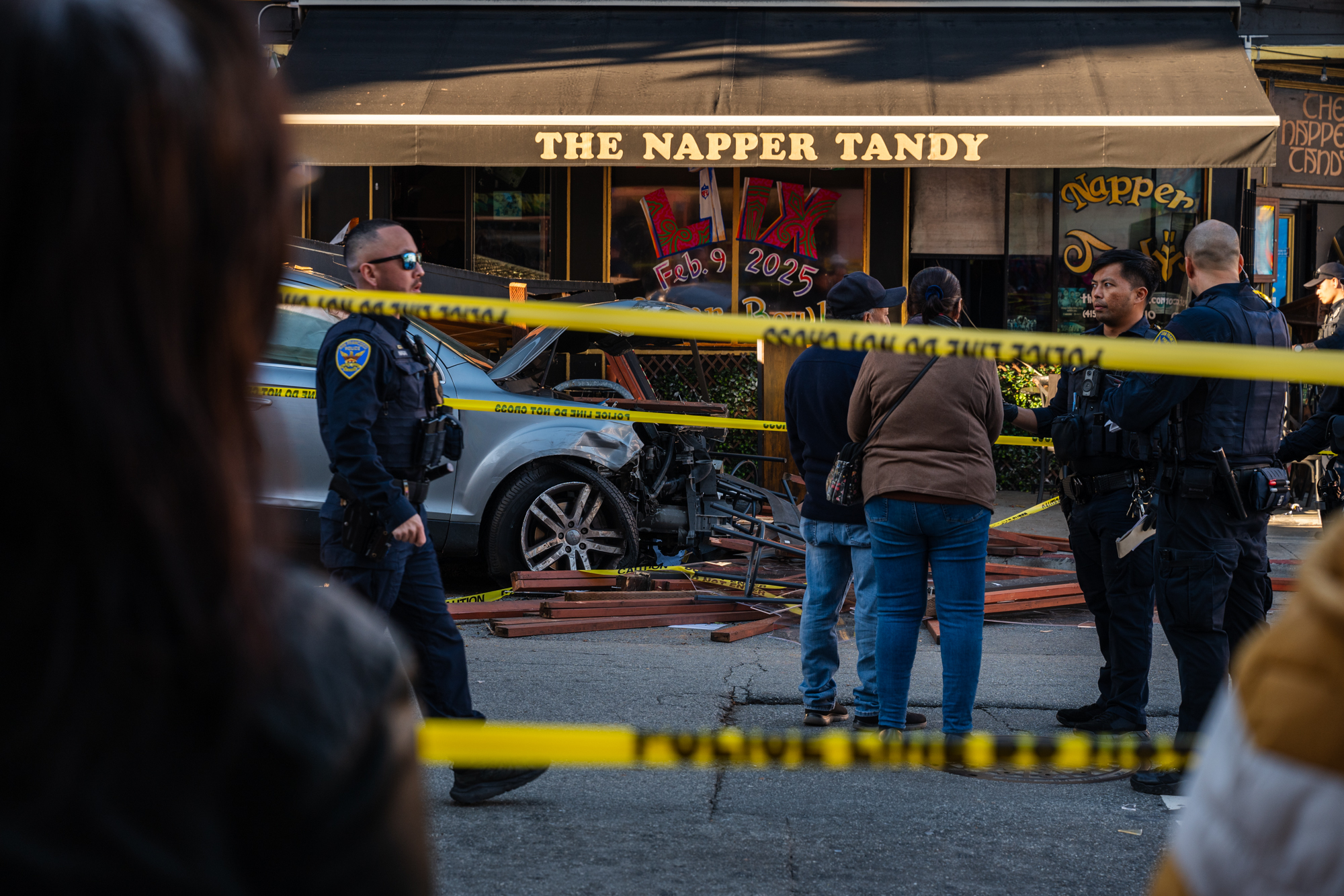 Police officers and bystanders at a car crash scene outside The Napper Tandy pub. Yellow caution tape blocks the area.