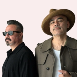 Two men posing against a plain background. One wears sunglasses and a black shirt; the other wears a brown coat and hat.
