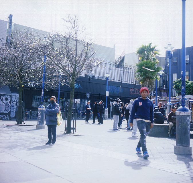 Urban scene with people gathered on a graffiti-covered street, some walking, others standing near leafless trees and a palm tree on a sunny day.