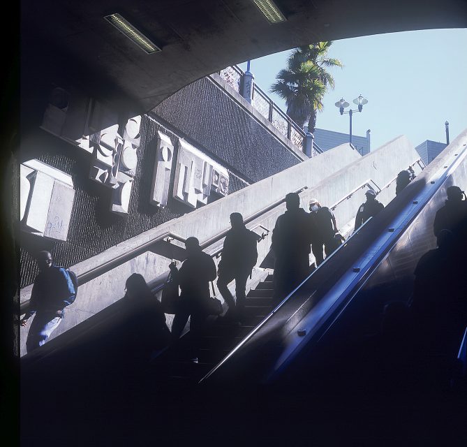 People on an escalator and stairs in a subway exit, silhouetted against daylight, with a palm tree and building visible above.