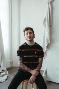 Person with tattoos sitting on a stool, wearing a black striped t-shirt, in a room with white walls and draped fabric.