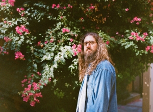 Man with long hair and beard in a denim jacket stands in front of a flowering bush with pink blooms.