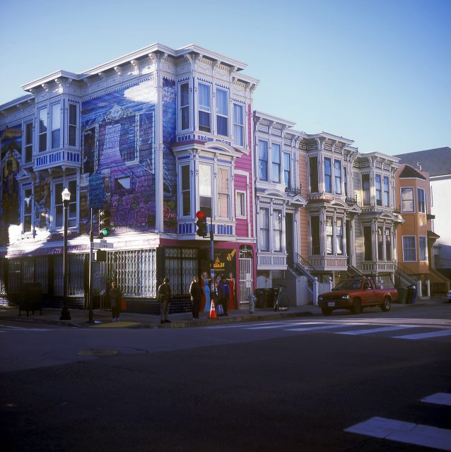 Colorful Victorian-style buildings with ornate facades line a street corner. A mural decorates one building. Pedestrians cross the street, and a red car is parked nearby.