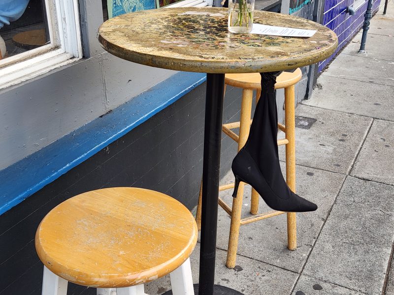 A round tabletop with a vase and napkin, supported by a metal pole, has a black high-heeled shoe hanging from it. Two wooden stools are placed around the table on a sidewalk.