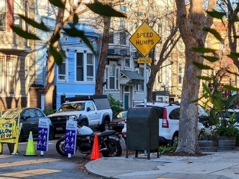 Residential street with traffic signs for speed humps, "Slow for Kids" notices, and parked cars. Trees and houses line the street.