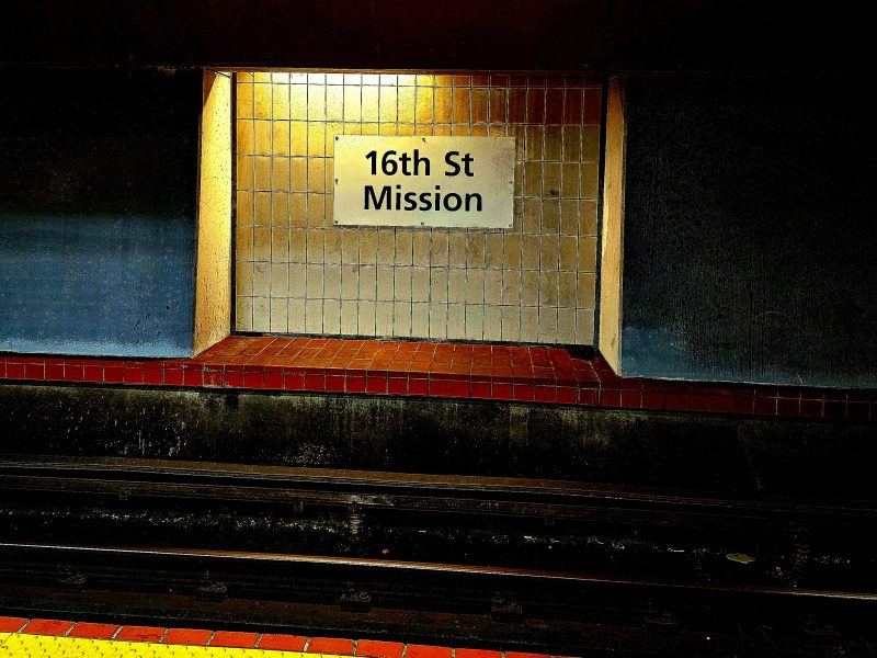Dimly lit subway station wall with a sign reading "16th St Mission" above empty tracks.