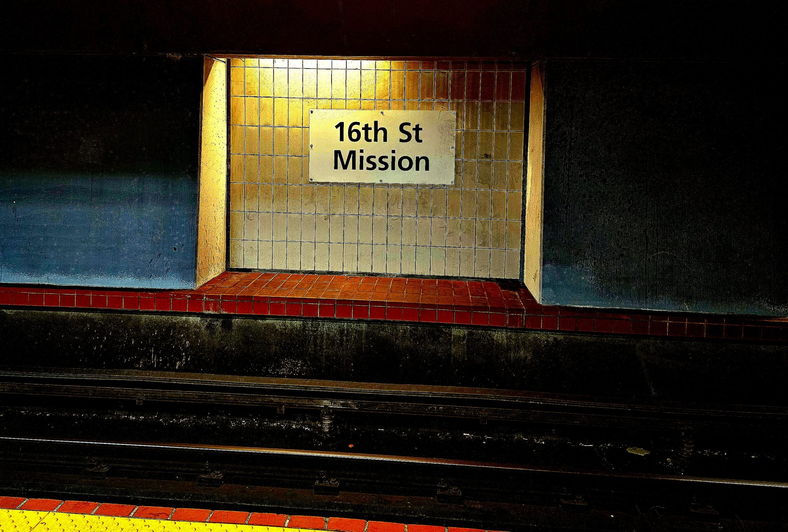 Dimly lit subway station wall with a sign reading "16th St Mission" above empty tracks.
