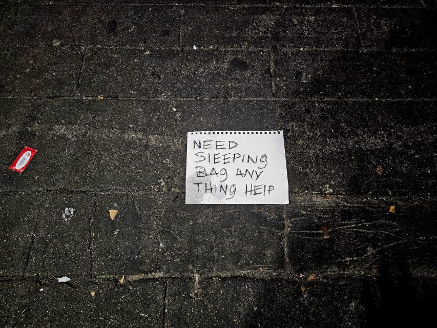 A handwritten sign on the ground reads, "Need sleeping bag any thing help," on a dark, worn pavement.