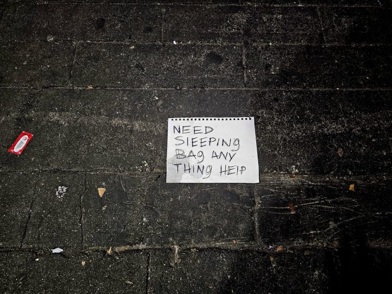 A handwritten sign on the ground reads, "Need sleeping bag any thing help," amid dark pavement with scattered litter nearby.