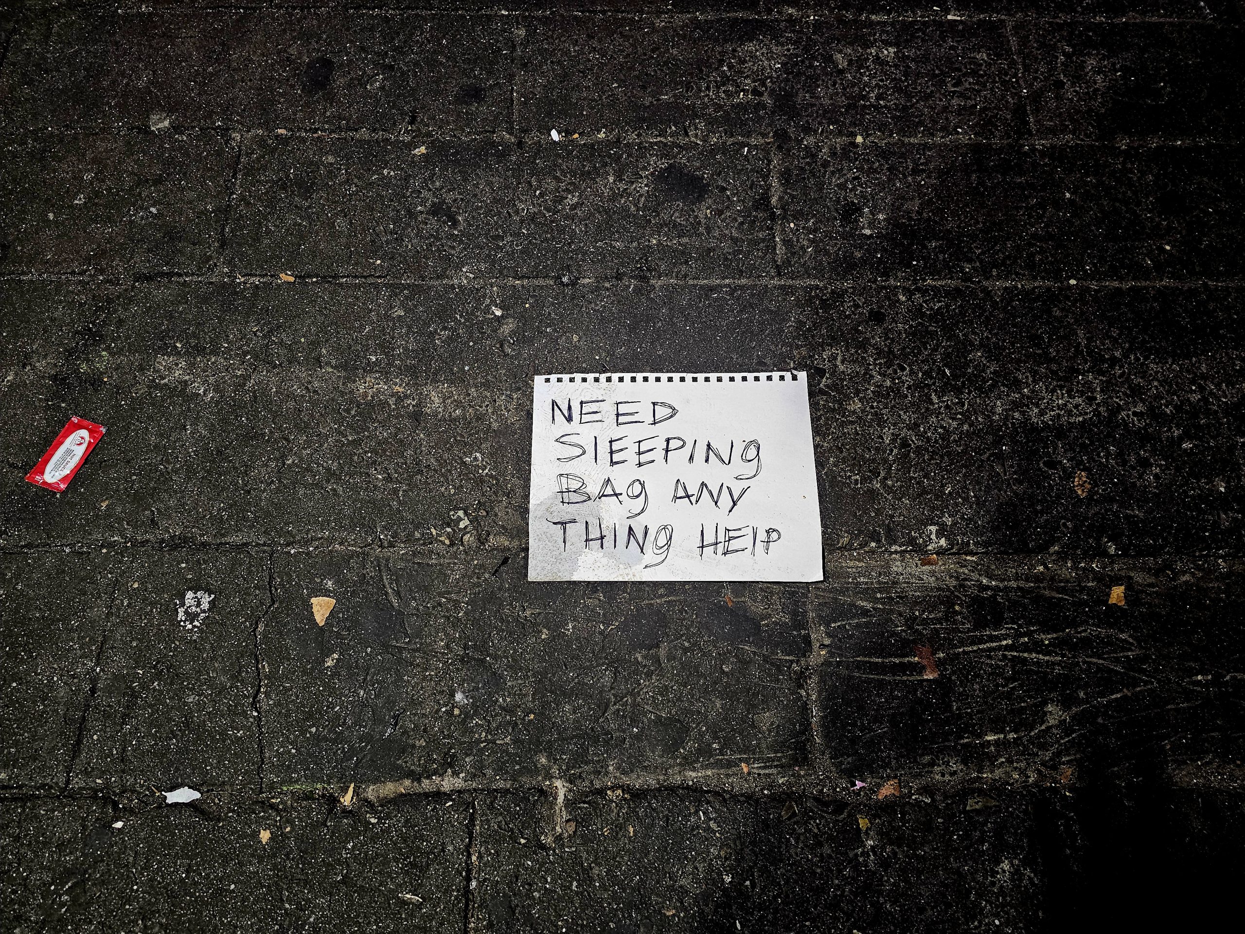 A handwritten sign on the ground reads, "Need sleeping bag any thing help," amid dark pavement with scattered litter nearby.