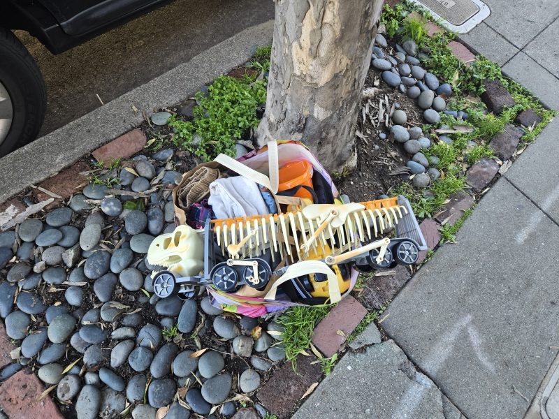 A pile of discarded toys and household items sits on the ground next to a tree trunk along a sidewalk.