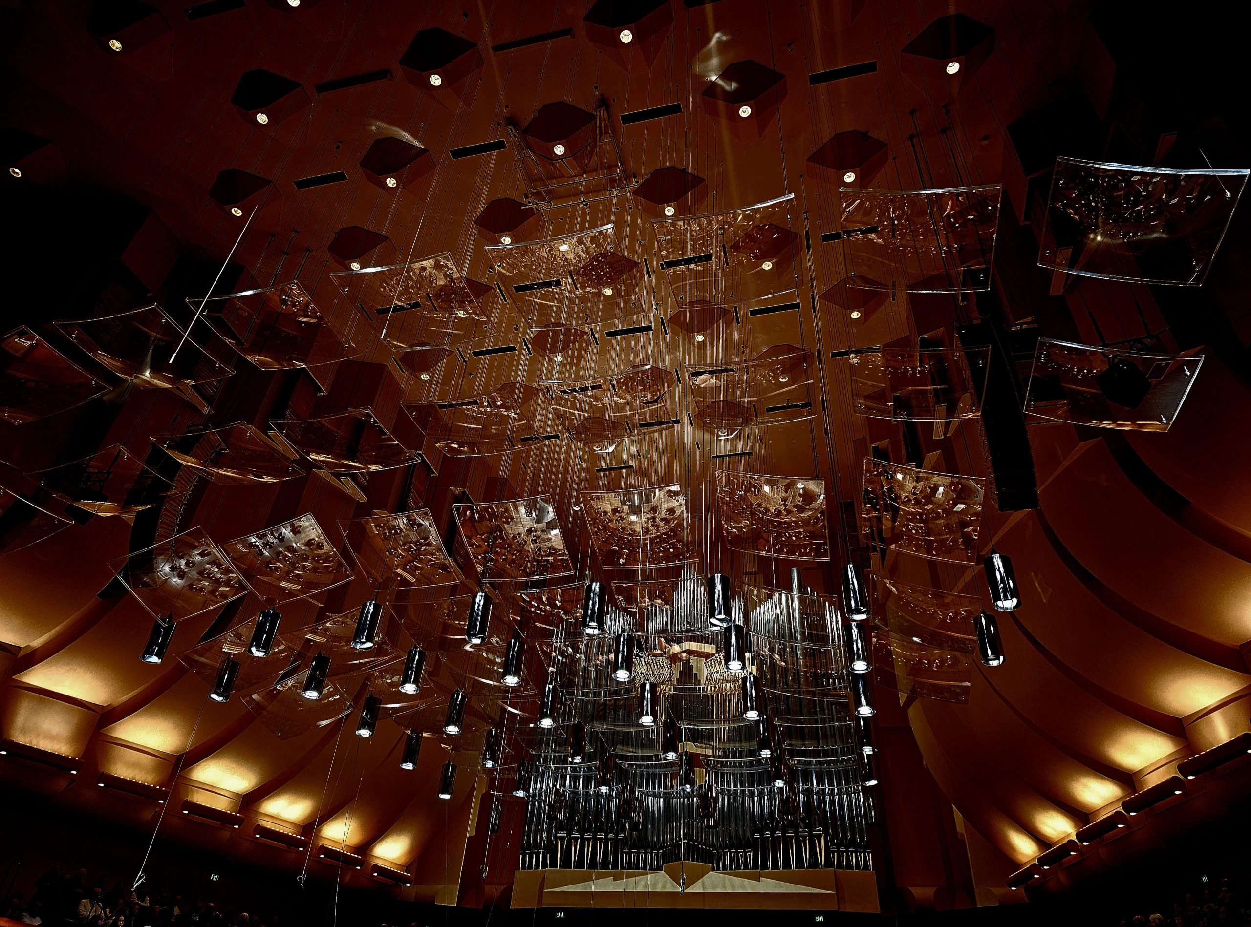 View of an ornate concert hall ceiling featuring suspended modern art installations and lighting fixtures, with an organ visible in the center.