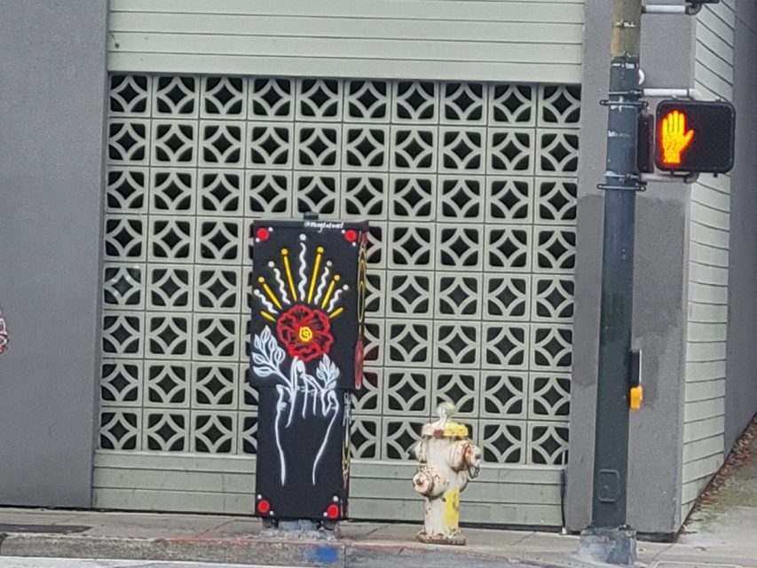 Utility box with colorful artwork of a red face surrounded by yellow rays and white floral designs, located near a fire hydrant and pedestrian crossing signal showing a red hand.