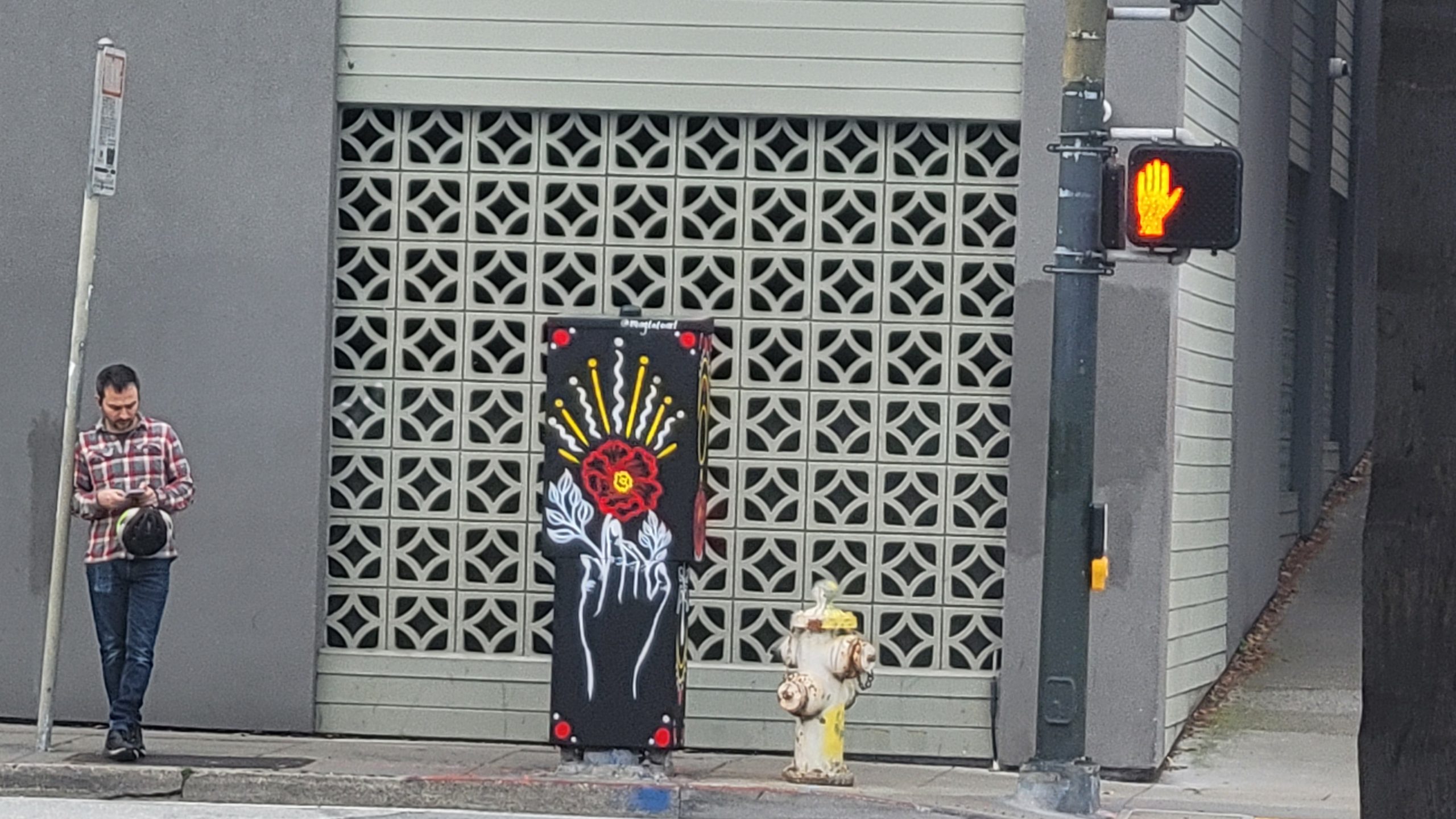 A man stands on the sidewalk holding a phone near a wall with graffiti of a red rose and white skeleton hand. A traffic light shows a red hand symbol.