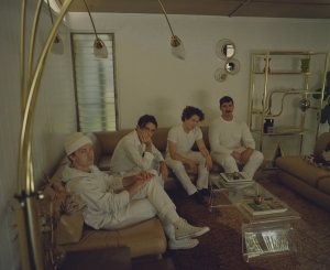 Four people in white outfits sit on a brown couch in a modern living room with a lamp and various decorations.