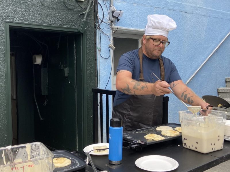 Person wearing a chef hat and apron making pancakes on a griddle outdoors. Containers and a blue bottle are on the table.
