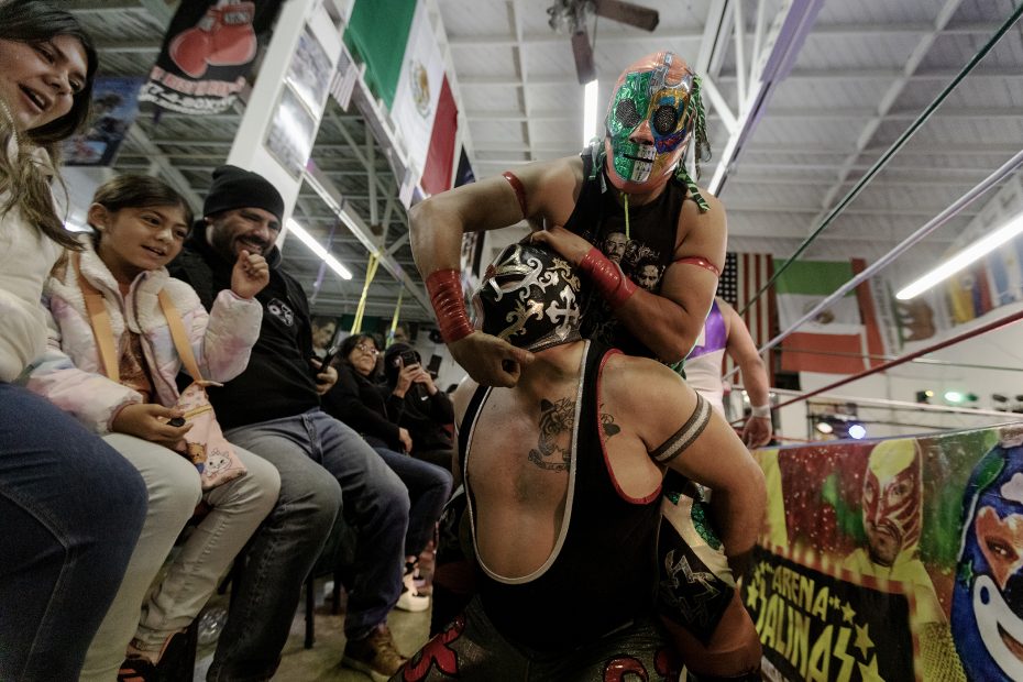 Two masked wrestlers engaged in a match near spectators, inside a wrestling ring arena decorated with flags.