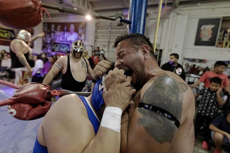 Two wrestlers, both in masks, are in a match. One is biting the hand of the other while a crowd watches.
