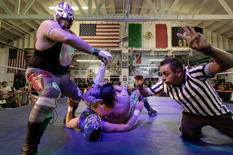 Two masked wrestlers in a ring, one pinning the other while a referee counts. American and Mexican flags are in the background.