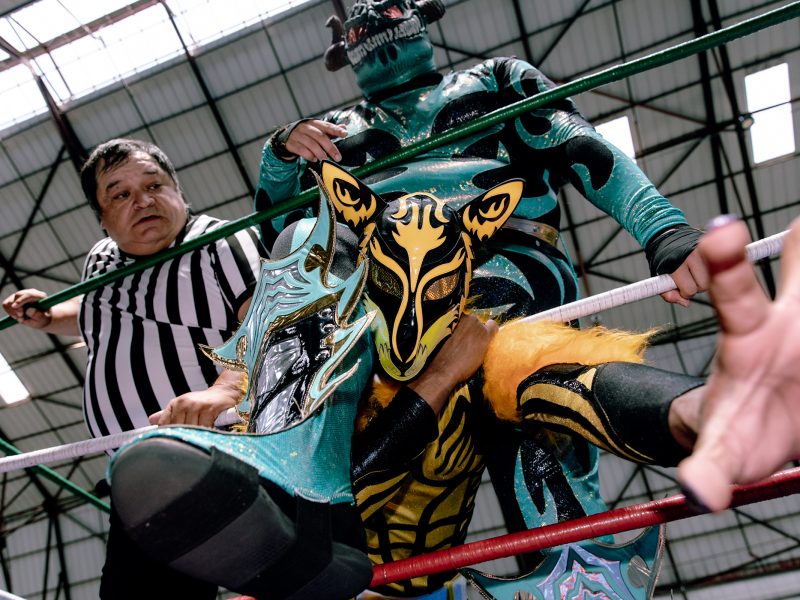 Wrestler in elaborate costume holds another wrestler's face with foot on ring ropes, while a referee looks on in a wrestling arena.