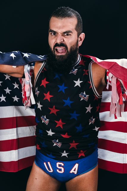 Person wearing star-patterned attire and USA-themed briefs, draped in an American flag, with a serious expression.