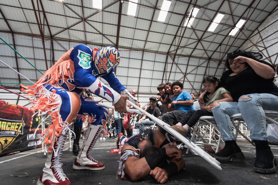 Wrestler in a blue costume attacks another wrestler on the ground with a chair, while spectators watch inside a gym with metal bleachers.