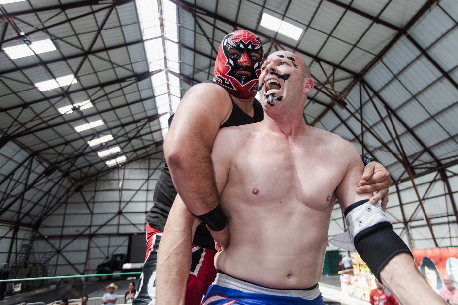 Two wrestlers in a warehouse-style venue. One wears a red mask and embraces the other, who is topless and smiling with black markings on his face. A wrestling ring is visible in the background.